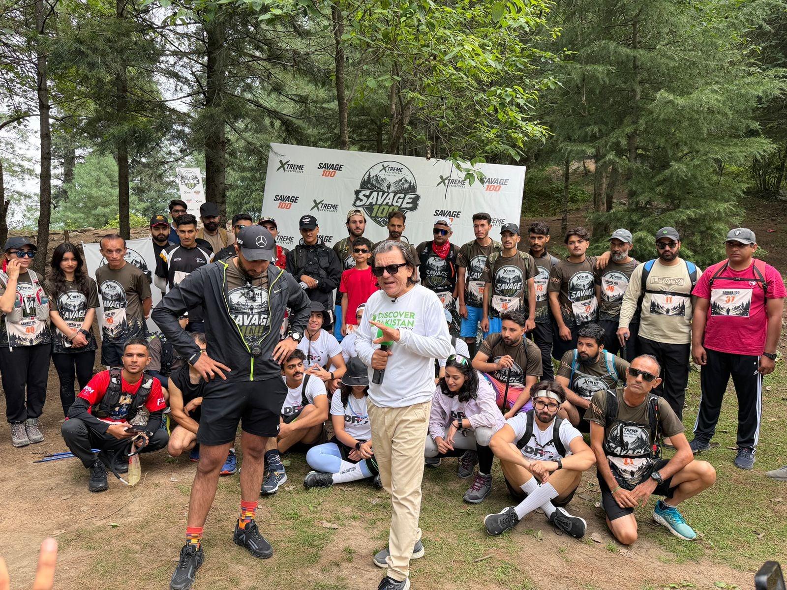 The Savage 100 field gathered in front of the event banner among cedar trees before the start, several dozen runners in race shirts standing and crouching together while a man in a white shirt speaks to them