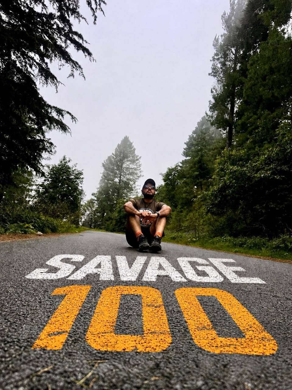 A runner crouched in the middle of a wet forest road with SAVAGE 100 painted across the tarmac in front of him in white and orange, cedars and mist closing in on both sides