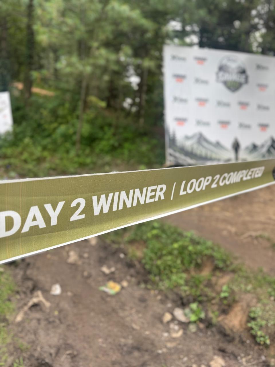 A close-up of an olive finish tape strung across a dirt trail, printed DAY 2 WINNER, LOOP 2 COMPLETED, with the branded backdrop out of focus behind it