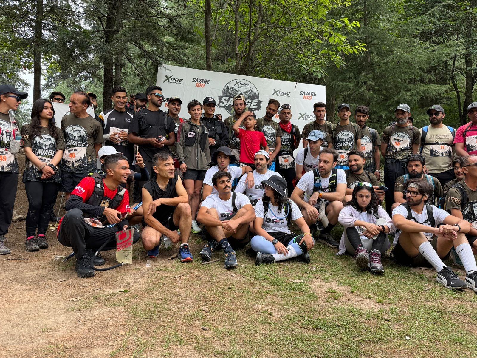 A wide group photograph of the Savage 100 entrants posed on a dirt clearing in front of the branded backdrop, runners standing in rows behind others kneeling and seated