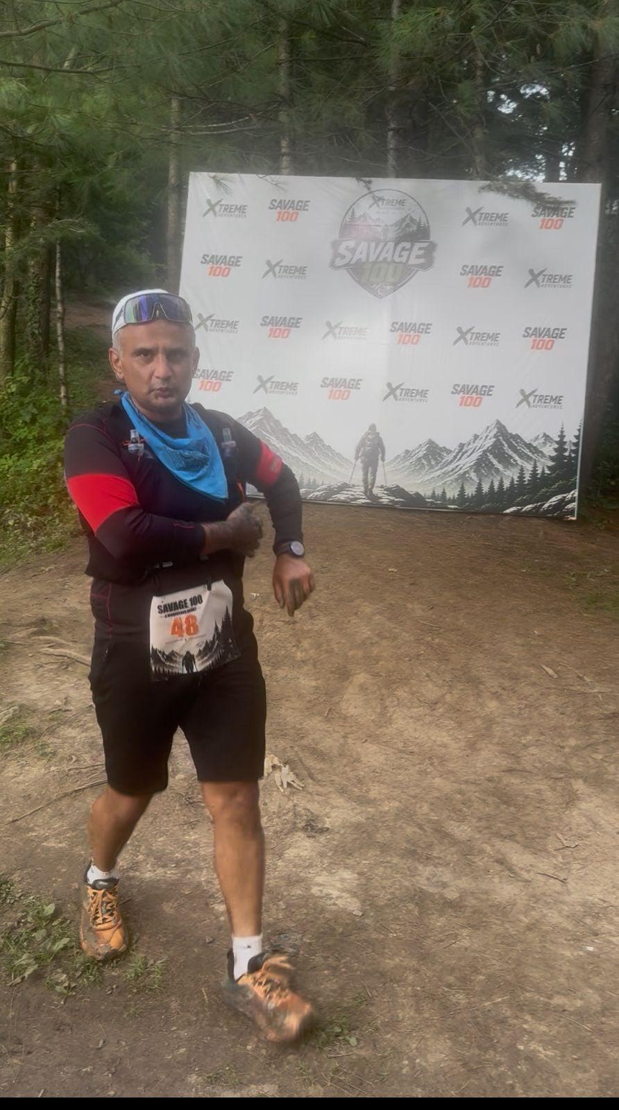 A runner wearing race number 48 walking a bare earth trail past the Savage 100 backdrop, the forest behind him lost in thick mist