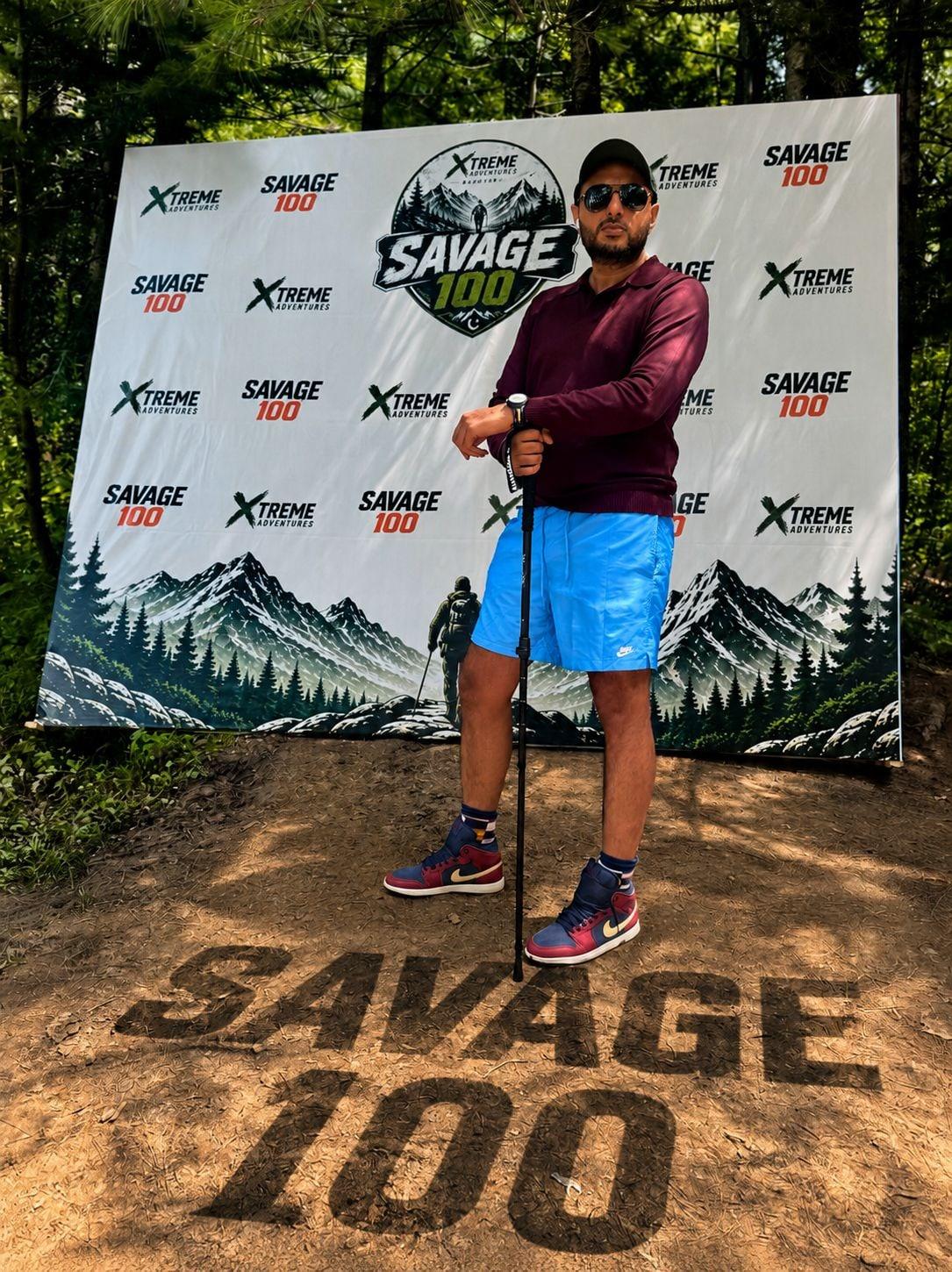 A runner in a maroon long-sleeved top and blue shorts standing with trekking poles in front of the Savage 100 backdrop, the words SAVAGE 100 scraped into the bare earth at his feet