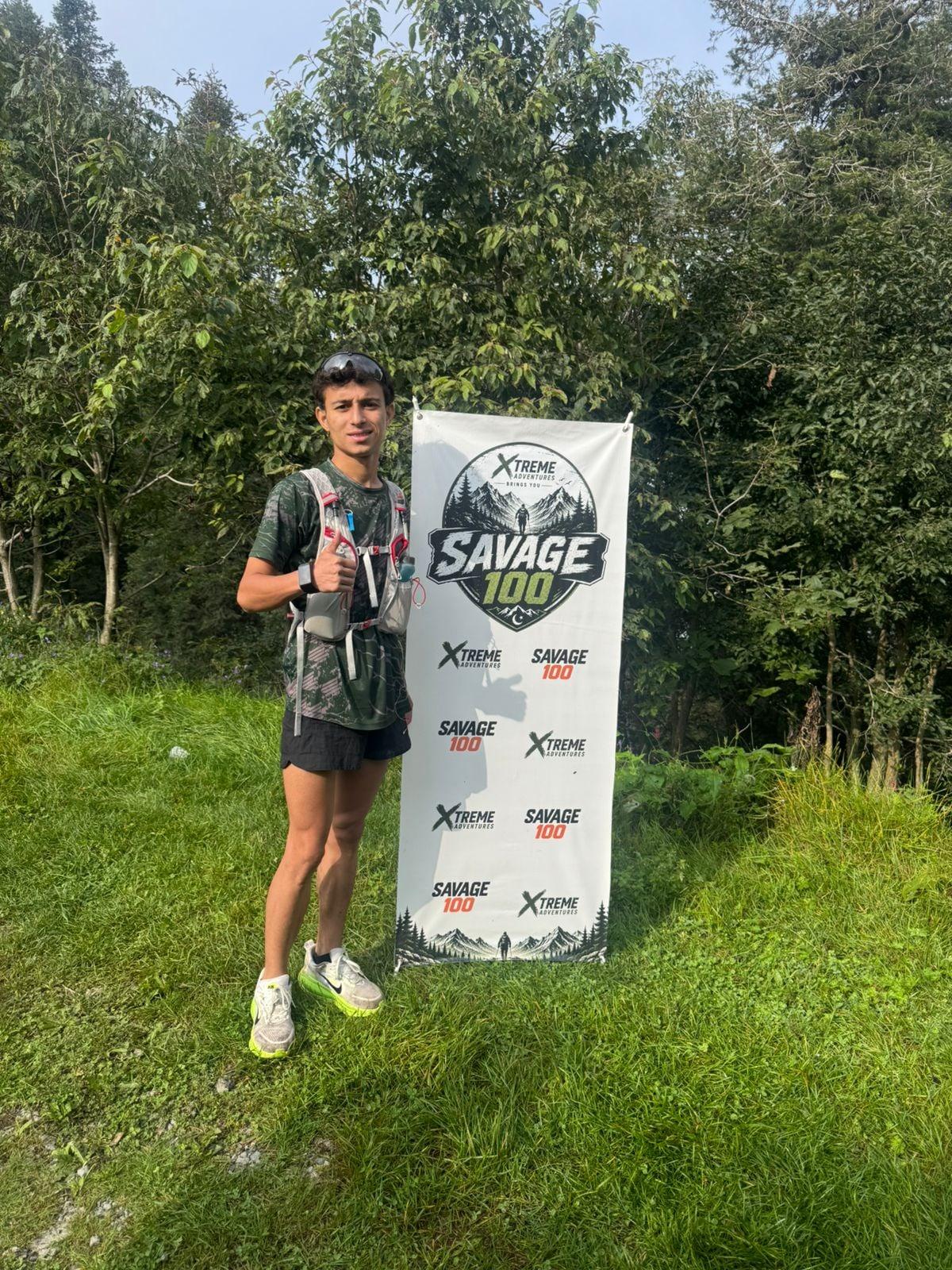 A young runner in a hydration vest standing on a grassy bank beside a tall vertical Savage 100 banner, dense green forest behind