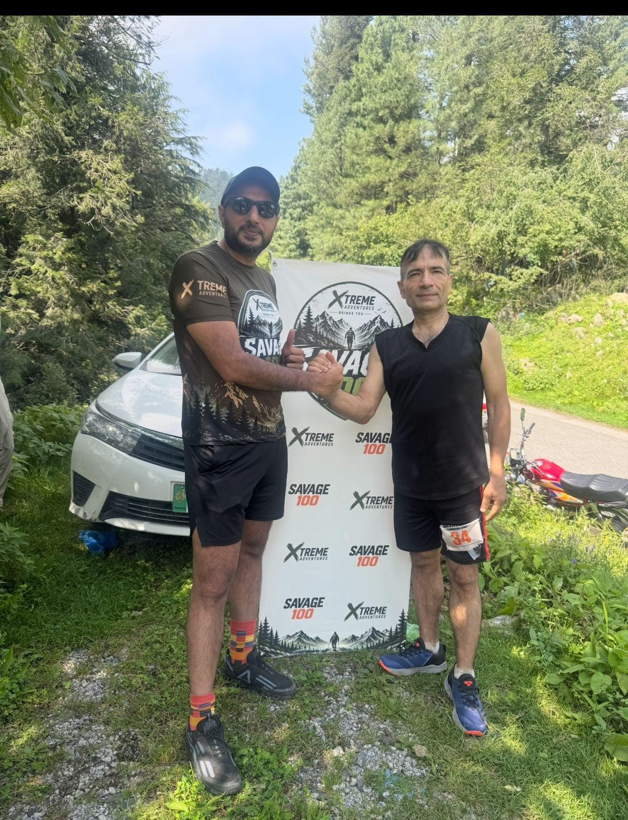 Two runners shaking hands in front of the Savage 100 backdrop after finishing, a white car and the forest road behind them