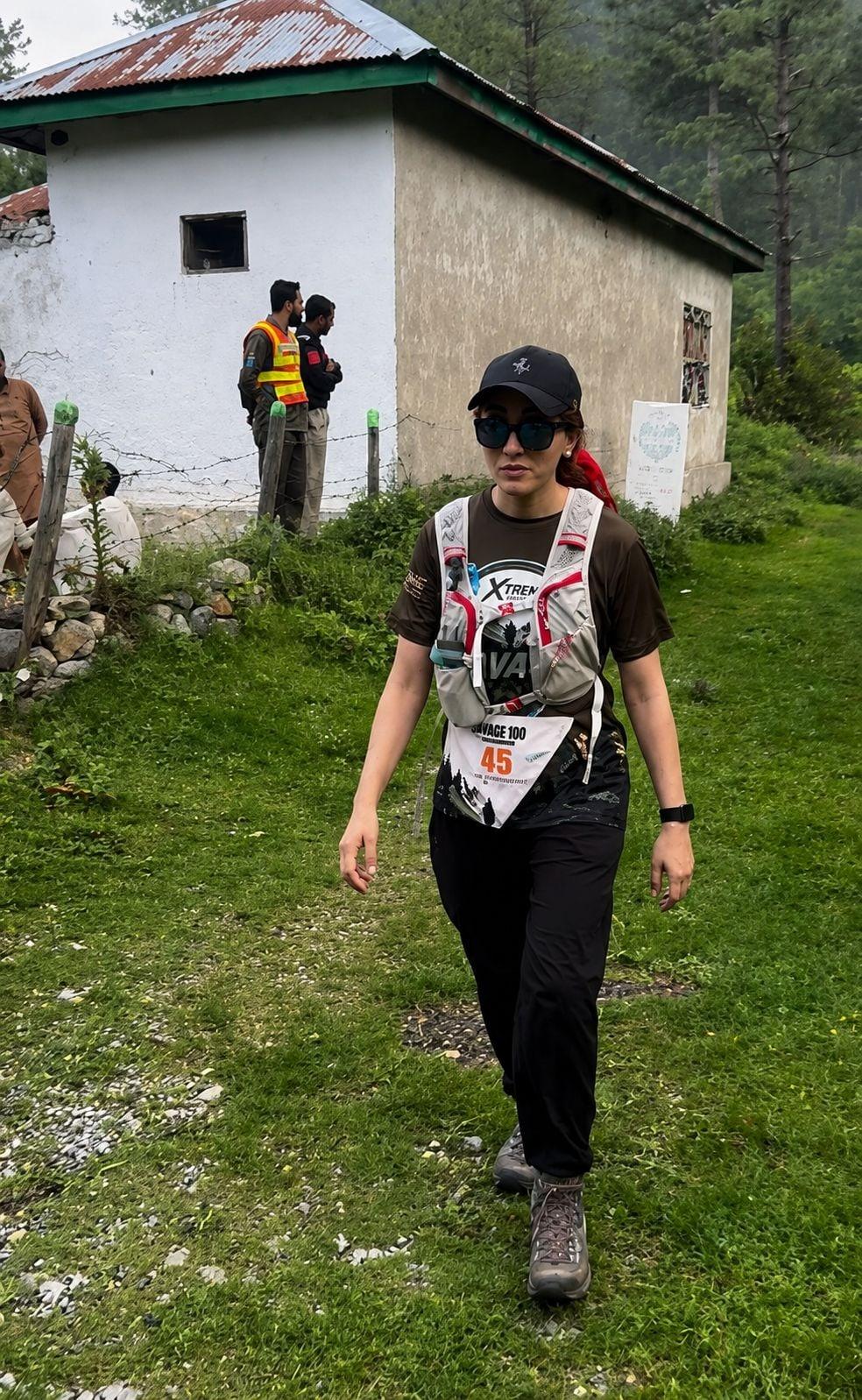 A woman wearing race number 45, a black cap, sunglasses and a hydration vest walking uphill on grass past a whitewashed stone building with a rusted tin roof