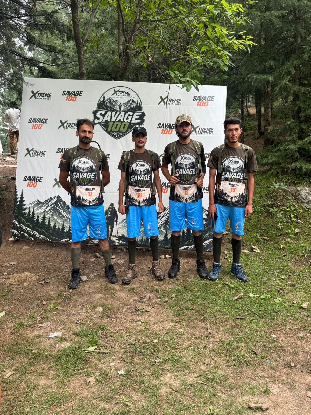 Four runners in matching Savage 100 shirts and blue shorts standing shoulder to shoulder in front of the branded backdrop in a forest clearing