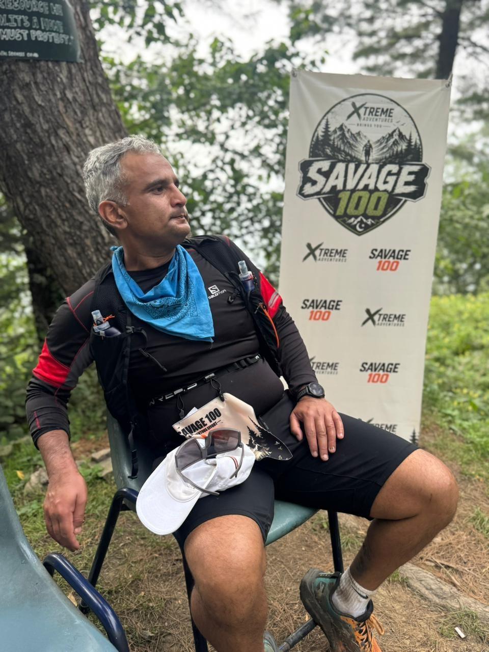 A grey-haired runner in a hydration vest sitting on a plastic chair beside the Savage 100 banner, cap held in his lap, looking up and away from the camera