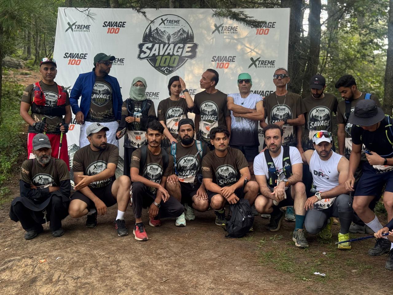 Around twenty runners in Savage 100 shirts posed in two rows in front of the branded backdrop in a forest clearing, one row standing and one crouching on the bare earth, hydration vests and trekking poles among them