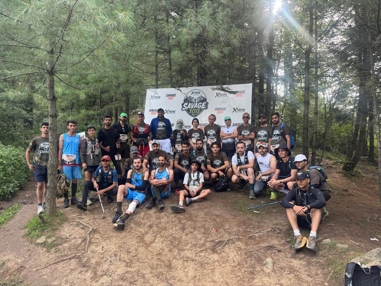 A large group of runners and organisers gathered on a dirt clearing under tall pines, the Savage 100 backdrop small behind them and sunlight breaking through the canopy