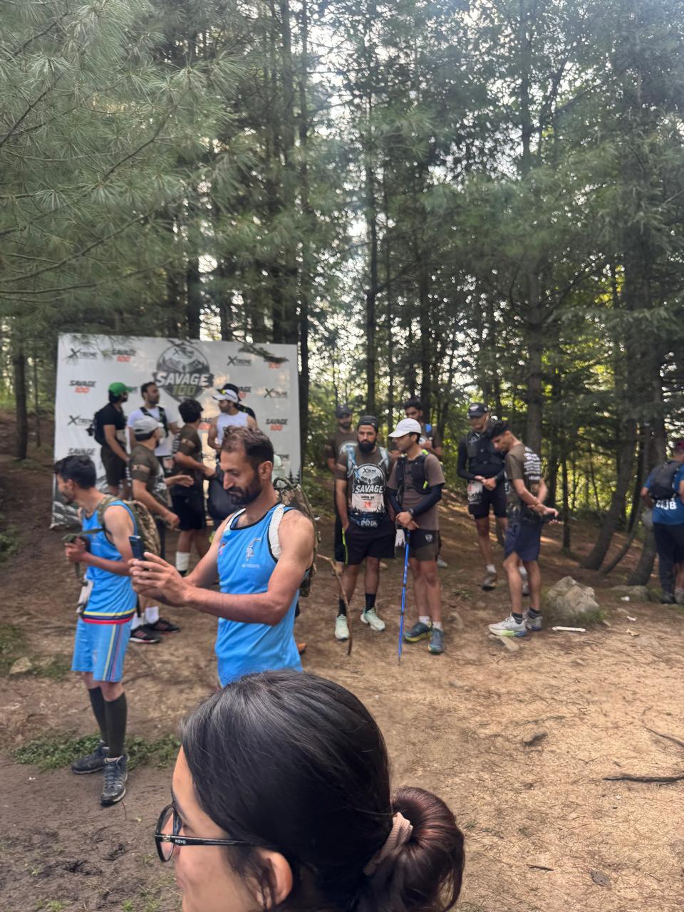 Runners waiting in the start area under the pines, two in pale blue vests talking in the middle of the frame, seen past the head of someone standing close to the camera