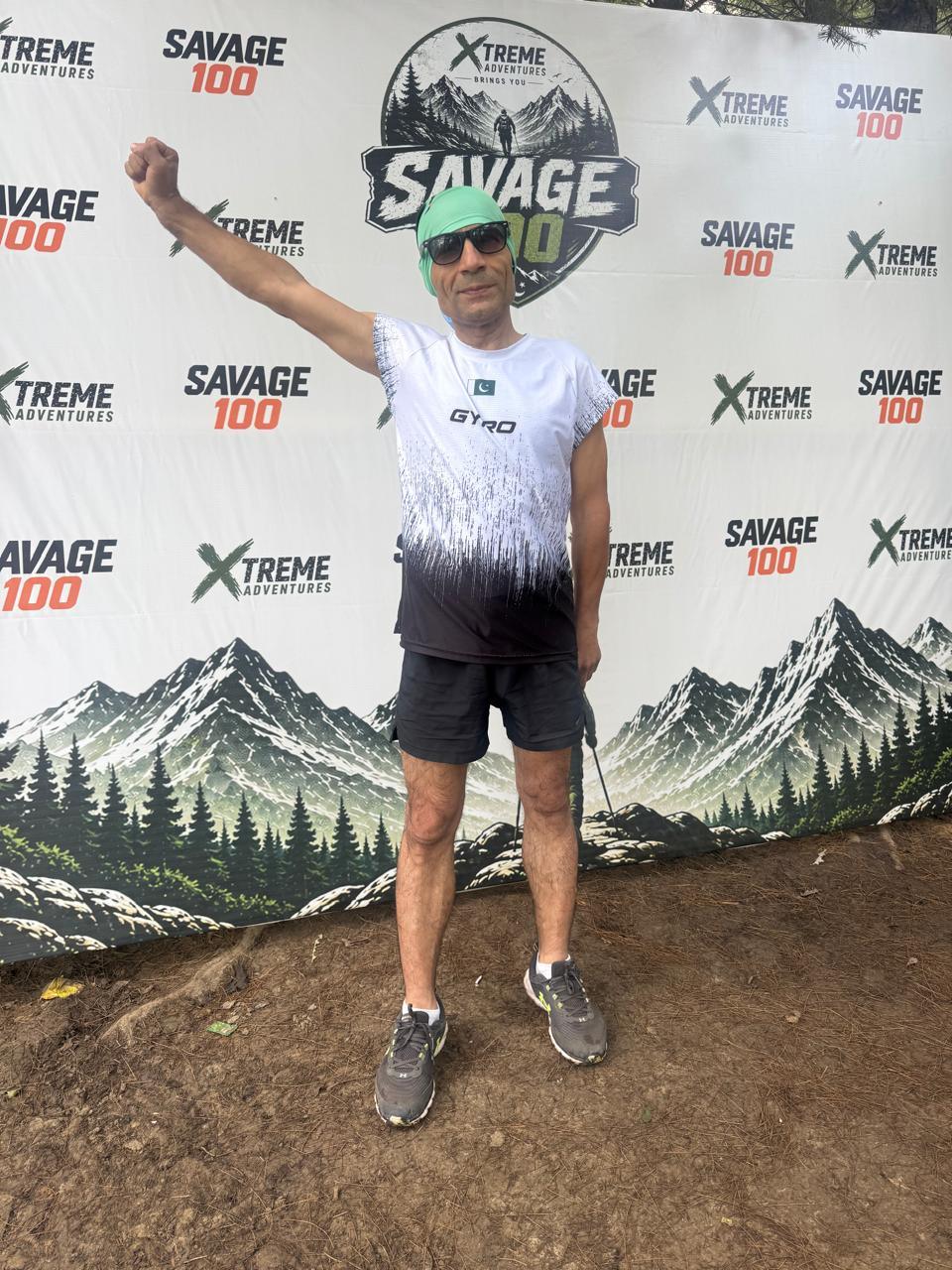 A grey-haired runner in a white and black gradient shirt and a green cap standing in front of the Savage 100 backdrop with one fist raised above his head