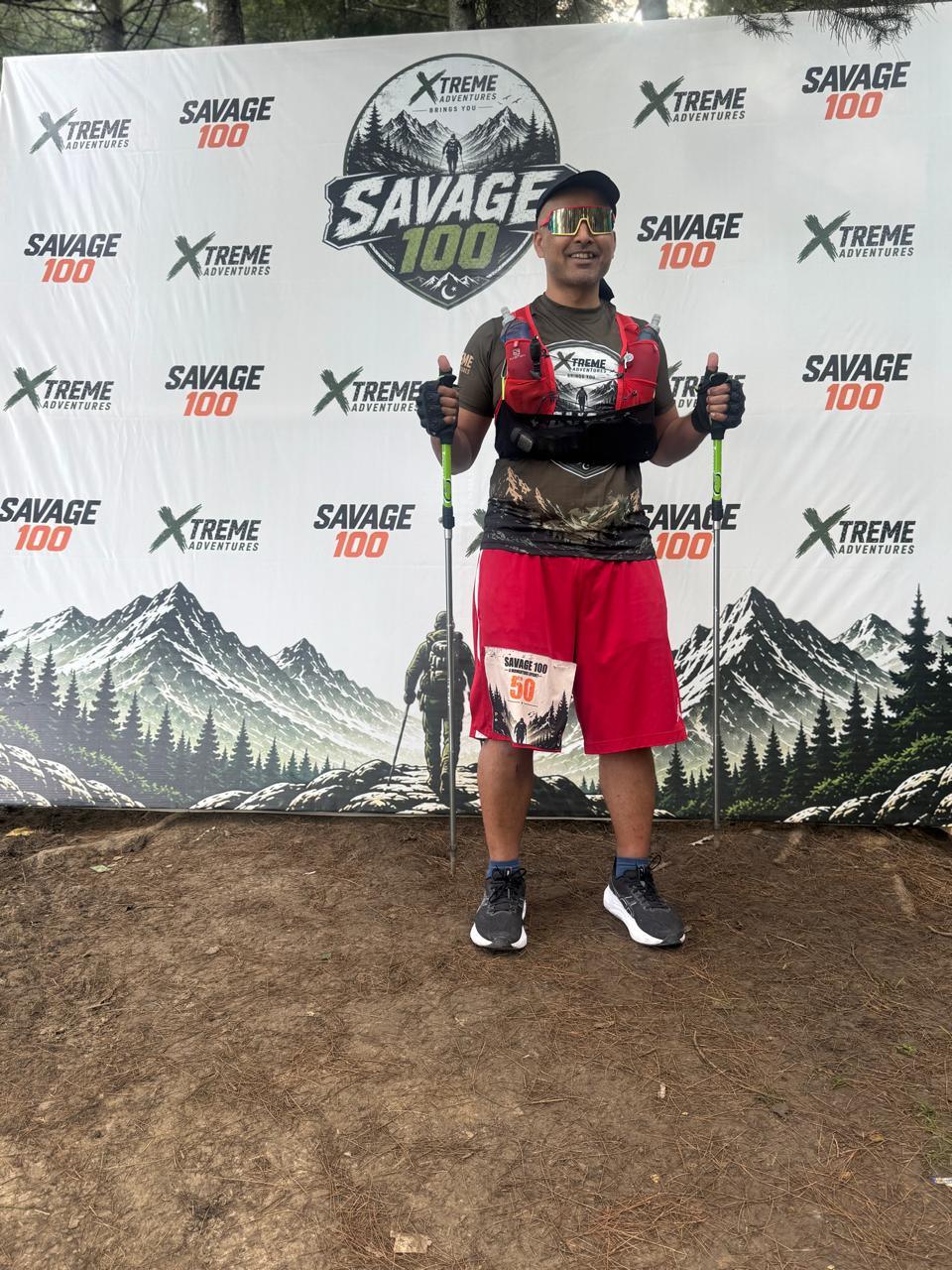 A runner in a camouflage-pattern shirt and red shorts standing squarely in front of the Savage 100 backdrop with a trekking pole in each hand and a race number pinned at his thigh