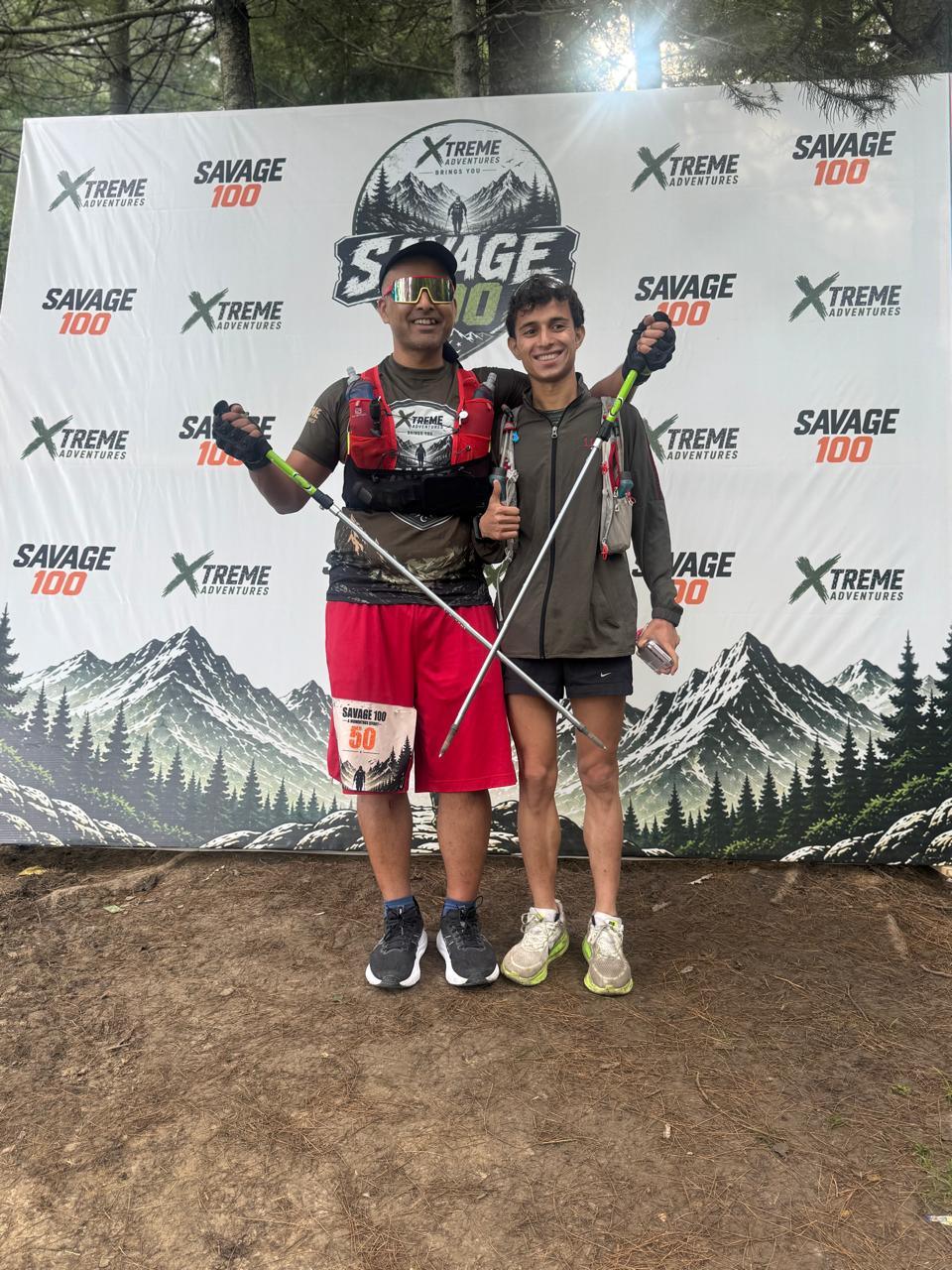 Two people in front of the Savage 100 backdrop, a runner in a red hydration vest holding trekking poles beside a younger companion in a grey top