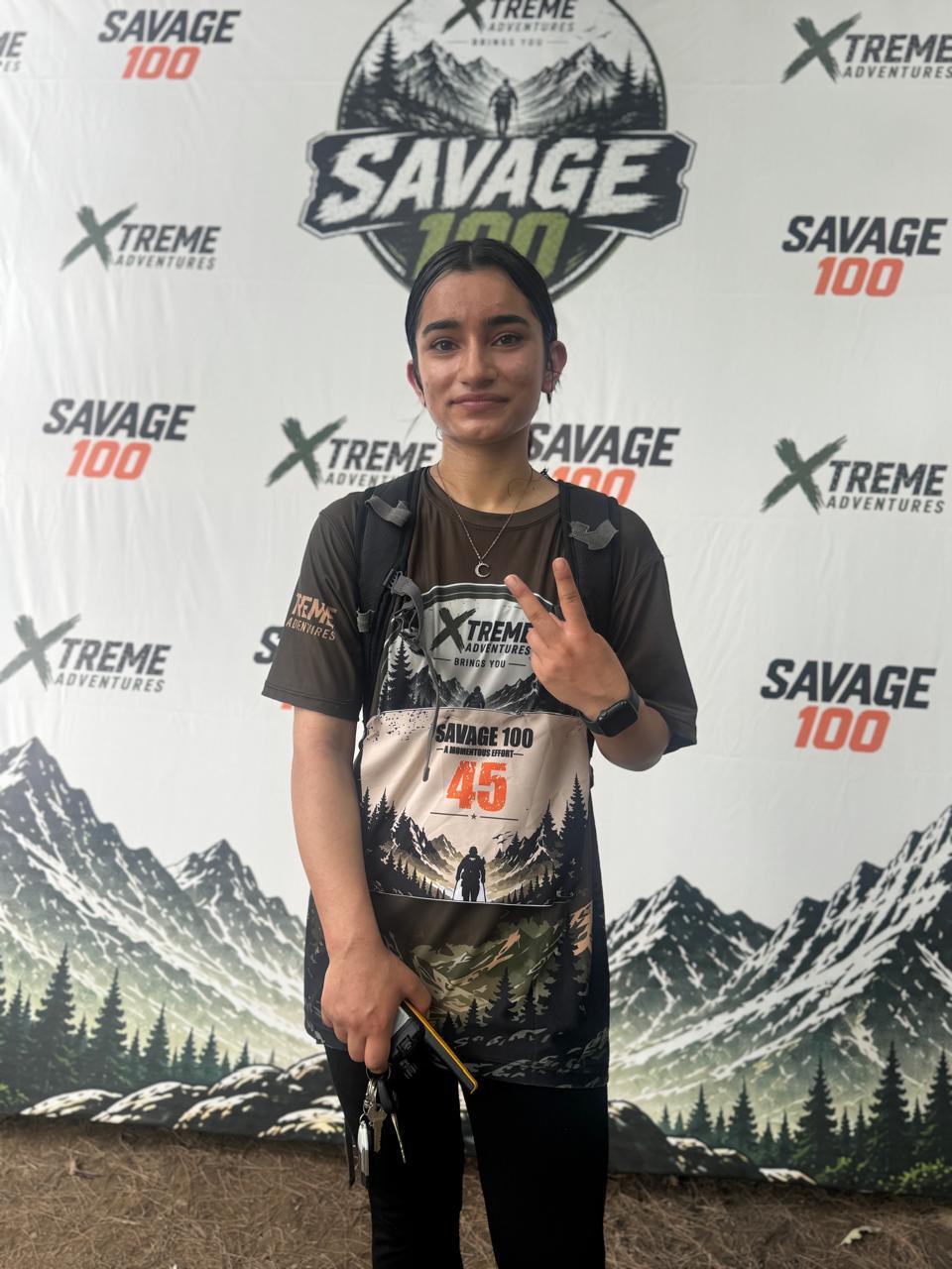 A woman wearing race number 45 over an Xtreme Adventures shirt standing in front of the Savage 100 backdrop, two fingers raised to the camera and a folded trekking pole in her other hand