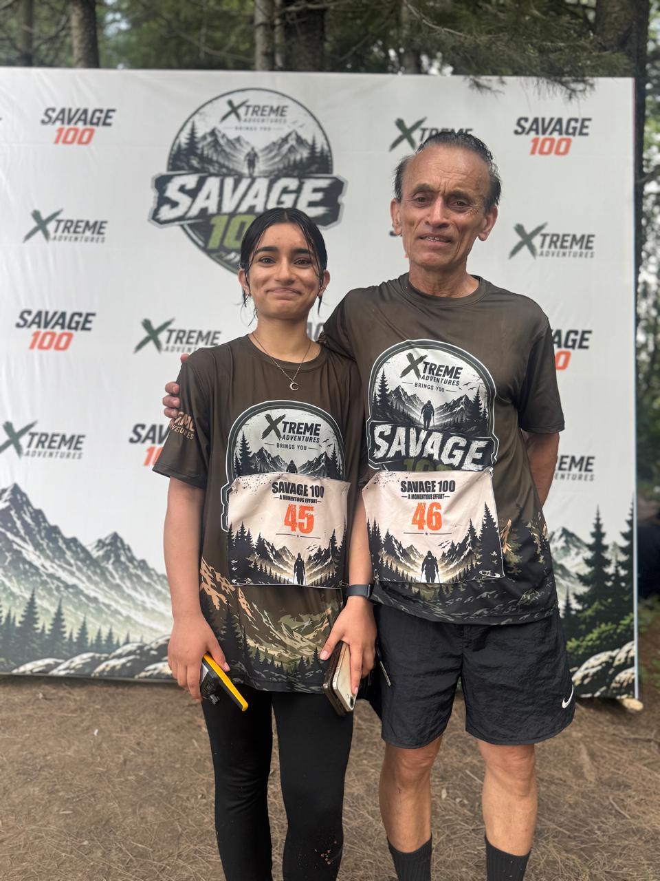 Two runners wearing numbers 45 and 46 standing side by side in front of the Savage 100 backdrop, a younger woman and an older man, both in Xtreme Adventures shirts