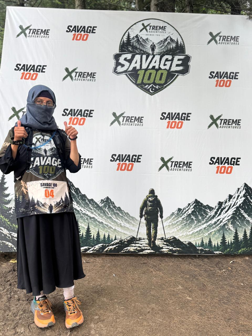 A runner wearing race number 04, a black headscarf and a face covering over an Xtreme Adventures shirt, standing in front of the Savage 100 backdrop with both thumbs raised