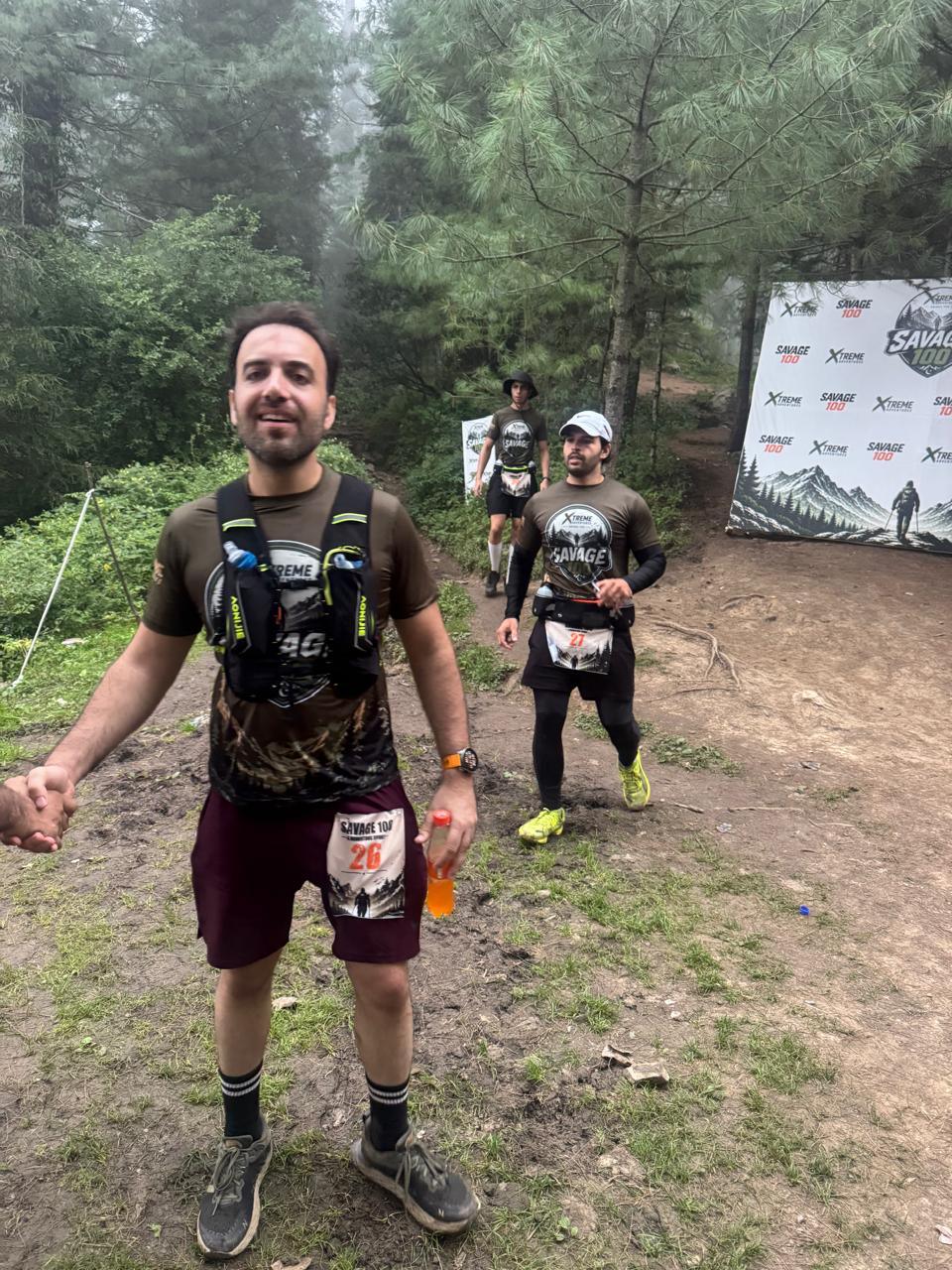 A runner in a maroon top coming up a bare earth trail towards the camera with a bottle in one hand, a second runner and the Savage 100 backdrop further up the slope behind him