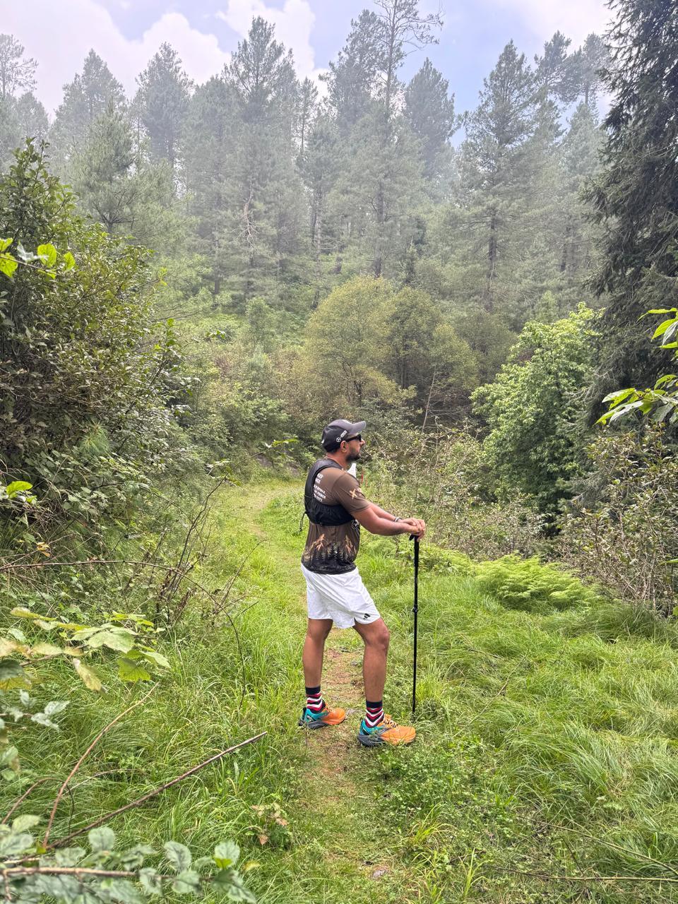 A runner in a dark Savage 100 shirt and white shorts working uphill through thick meadow grass with one pole planted, forest closing in on both sides