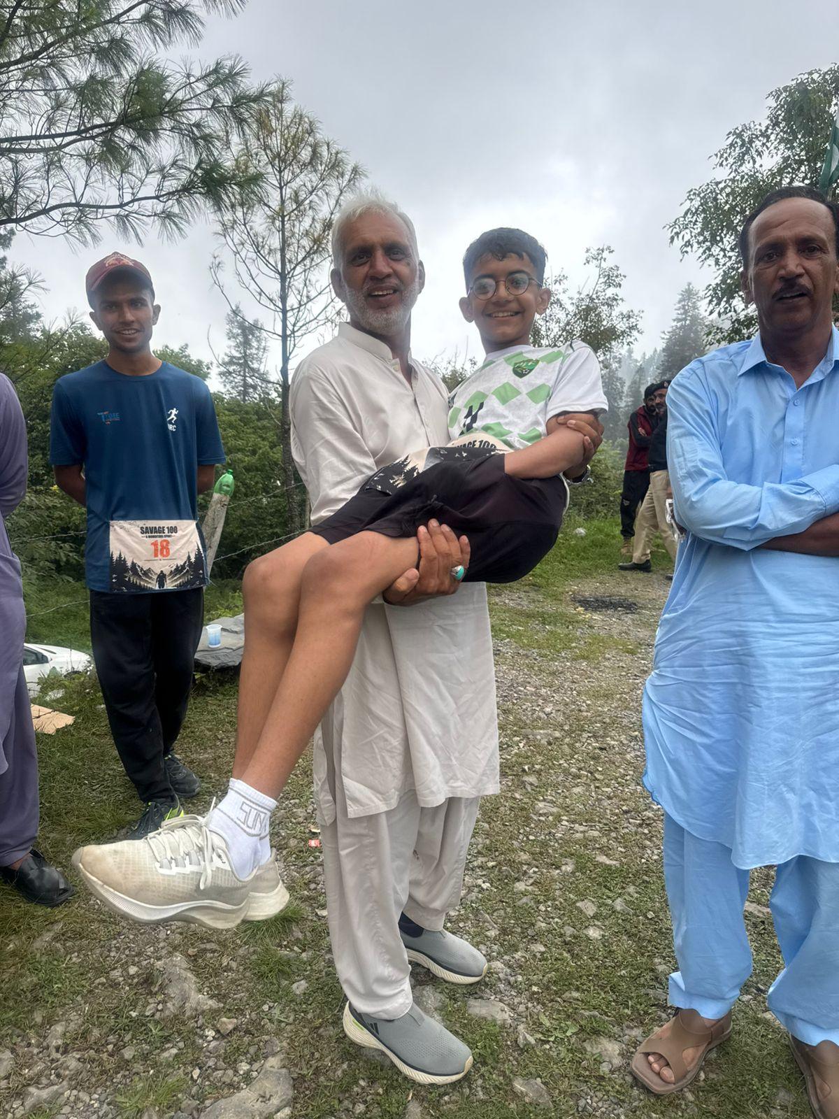 An older man in a white shalwar kameez carrying a laughing boy in his arms on a grass verge, a runner wearing number 18 standing beside them and a man in pale blue at the edge of the frame