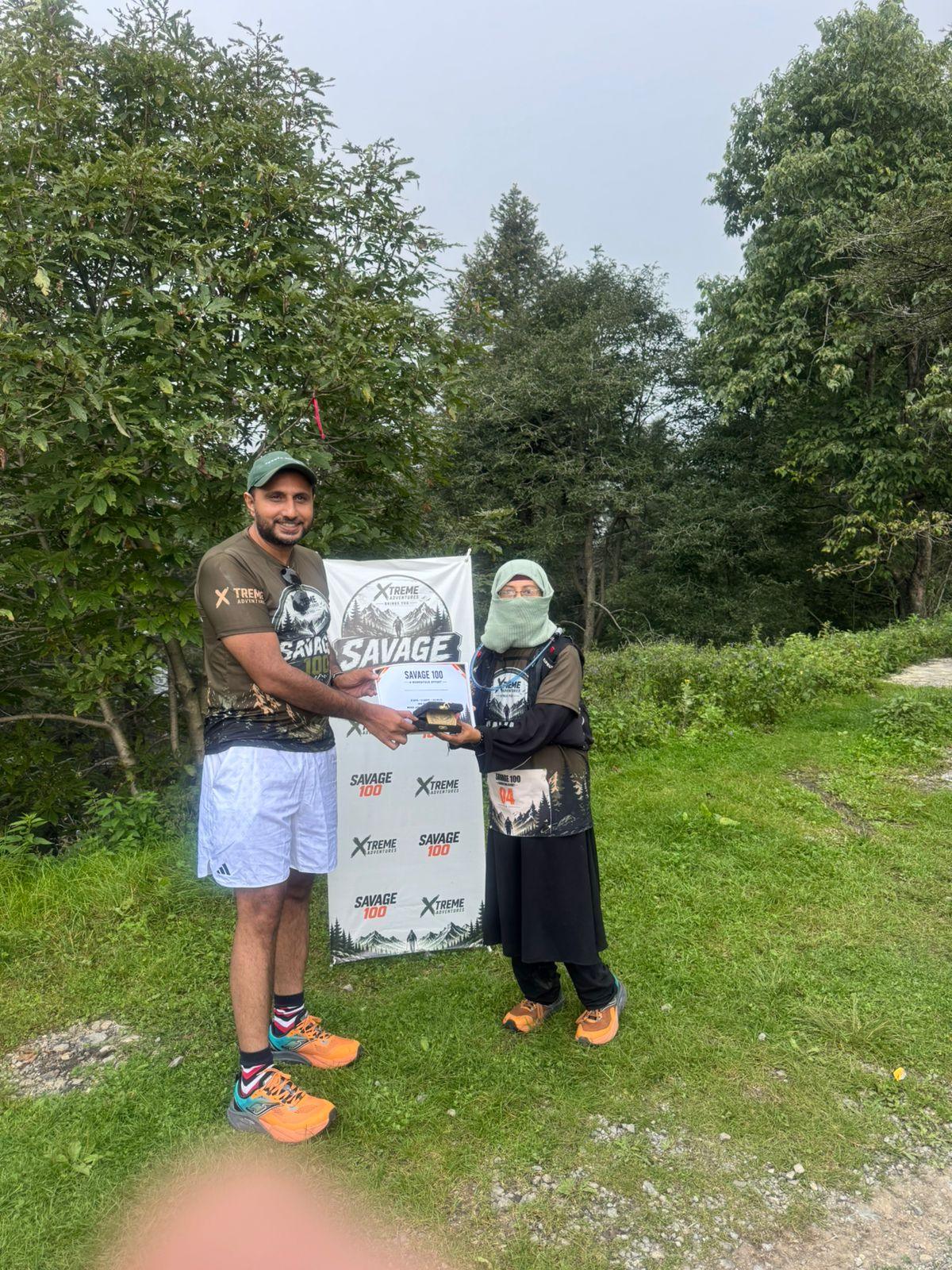 An organiser in a dark Savage 100 shirt handing a certificate to a woman in a black abaya and headscarf on a grass slope, the pair holding a small branded banner between them