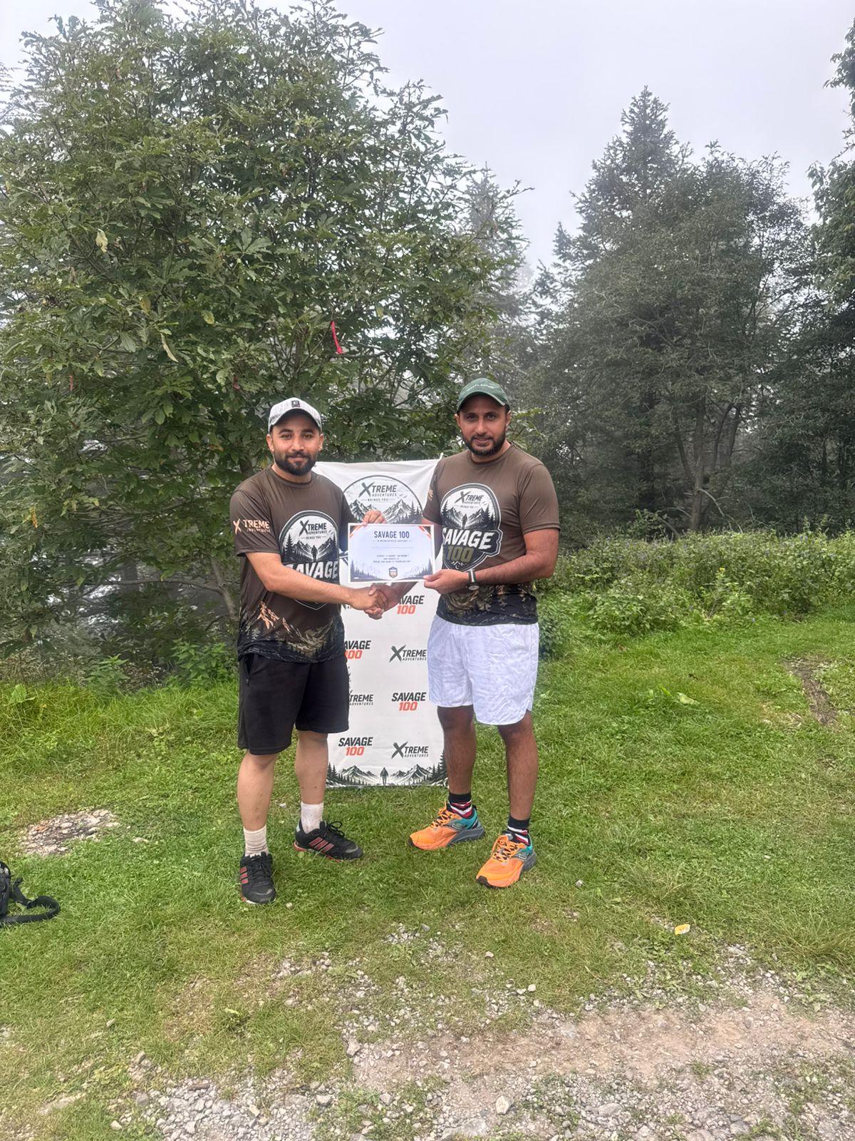 The organiser handing a certificate to a finisher in an olive shirt and white shorts, both holding the branded banner between them on the grass