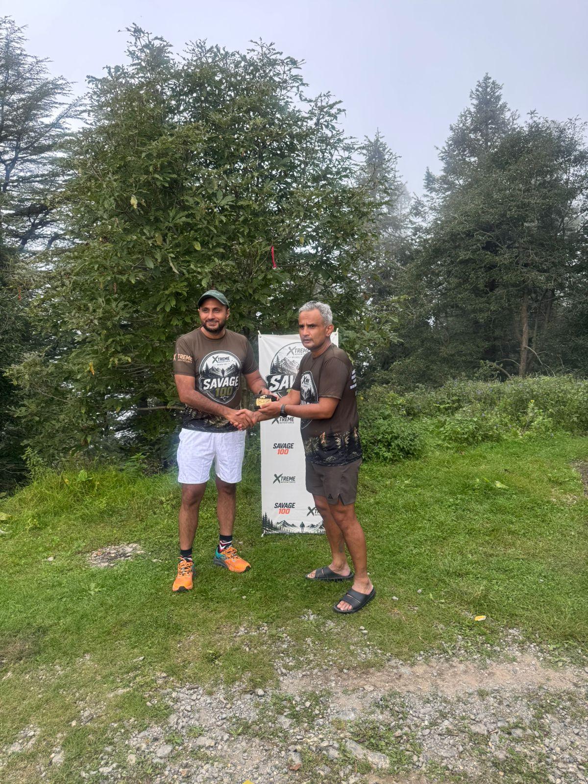 The organiser presenting a certificate to an older finisher in a dark shirt, grey shorts and sandals, forest behind them