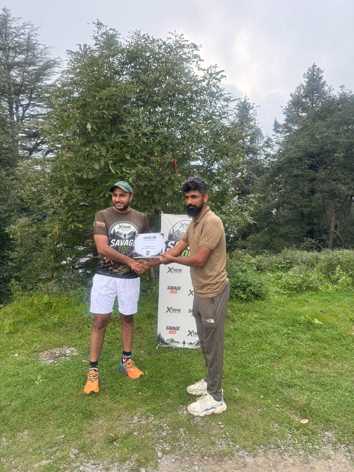 The organiser handing a certificate to a bearded finisher in an olive shirt and dark trousers, the branded banner held between them