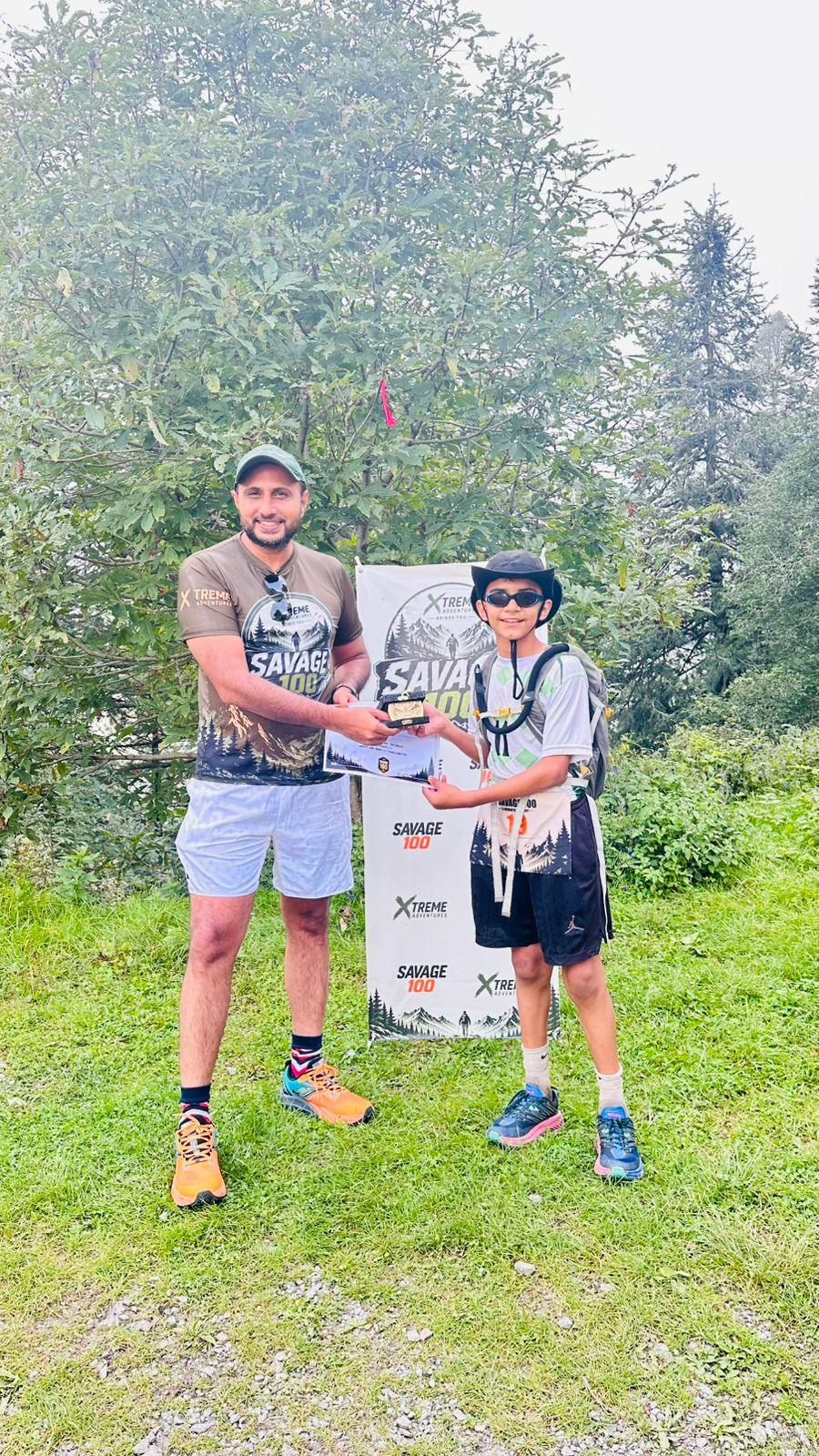 The organiser presenting a certificate to a young finisher in a black cap and sunglasses, both looking at the camera over the branded banner
