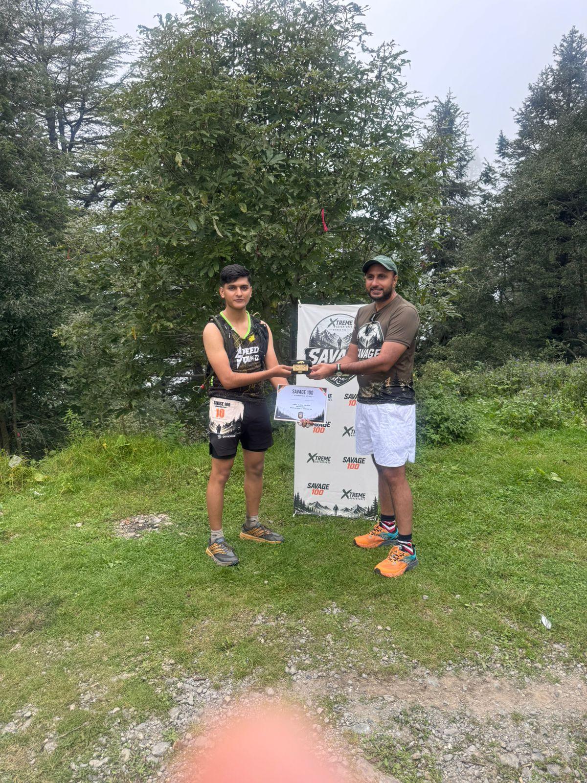 The organiser handing a certificate to a young runner in a black and green vest still wearing a race number