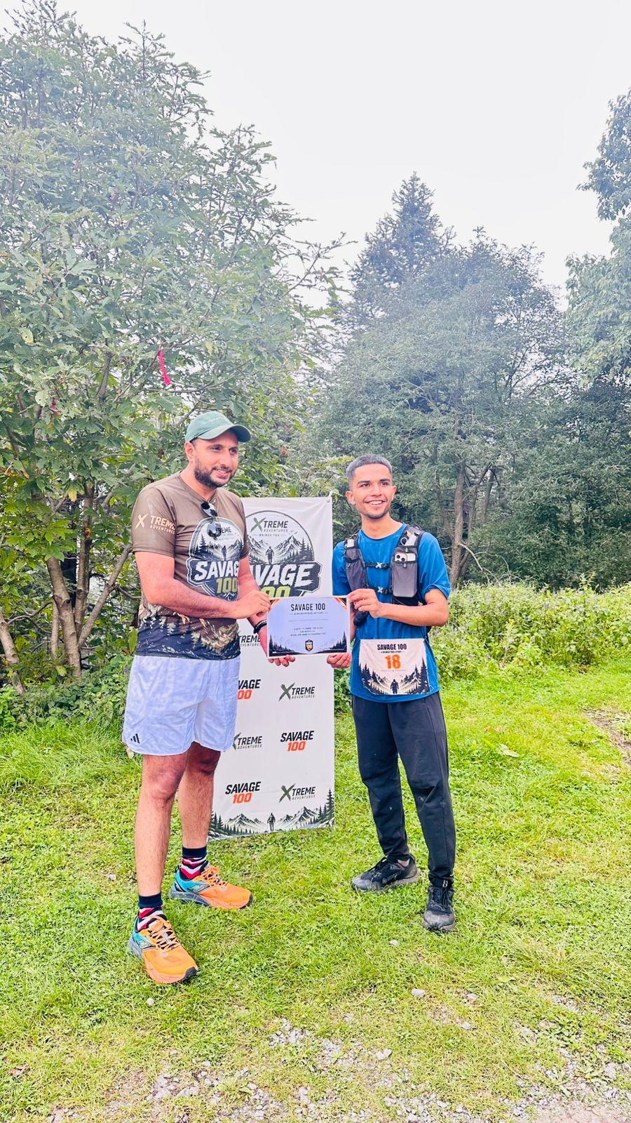 The organiser presenting a certificate to a finisher in a blue top and hydration vest on the grass slope