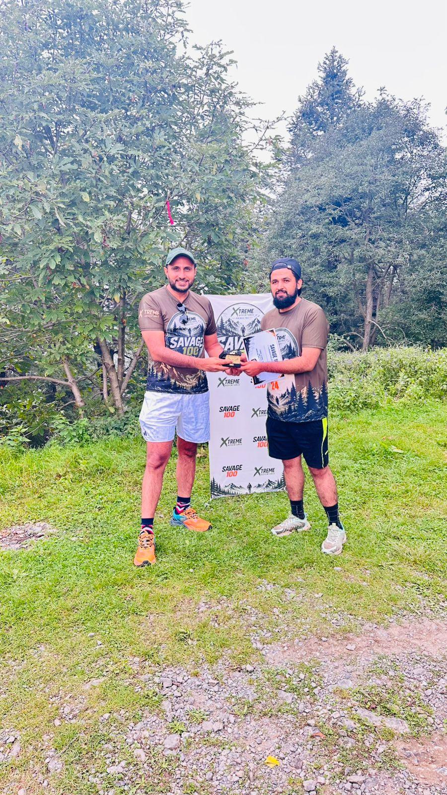 The organiser and a finisher in a Savage 100 shirt and black shorts holding a certificate and a small trophy between them