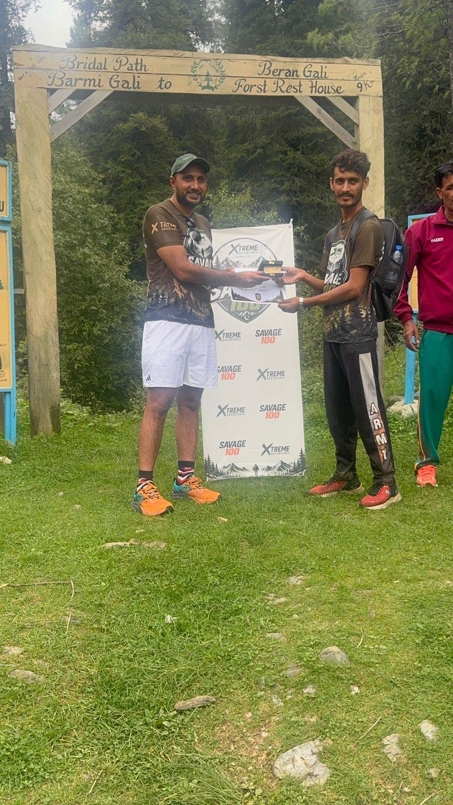 A certificate presented under a painted wooden gate reading Bridal Path, Barmi Gali to Beran Gali Forest Rest House, a marshal in a maroon top and green trousers standing to one side with a clipboard