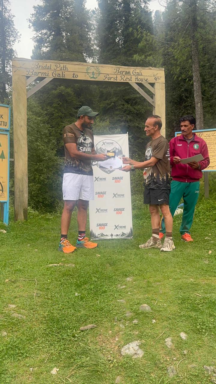The organiser handing a certificate to an older finisher in a dark shirt and shorts beneath the wooden gate, the marshal with the clipboard beside them