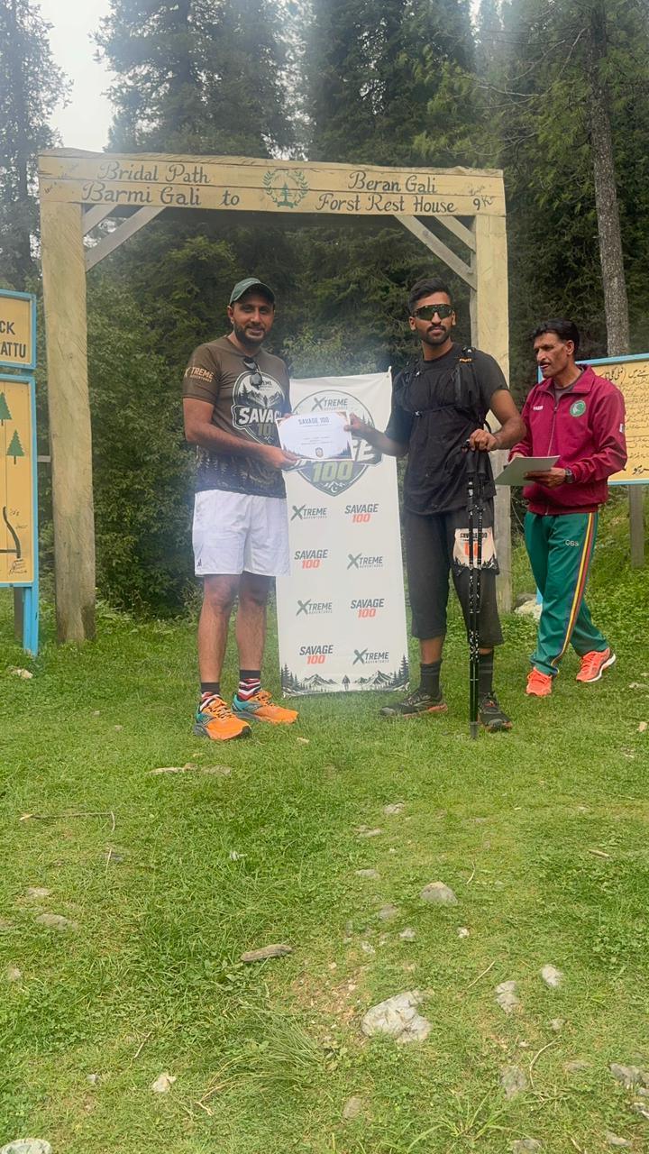 A tall finisher in a black long-sleeved top and boots receiving a certificate under the gate, forest and cloud behind