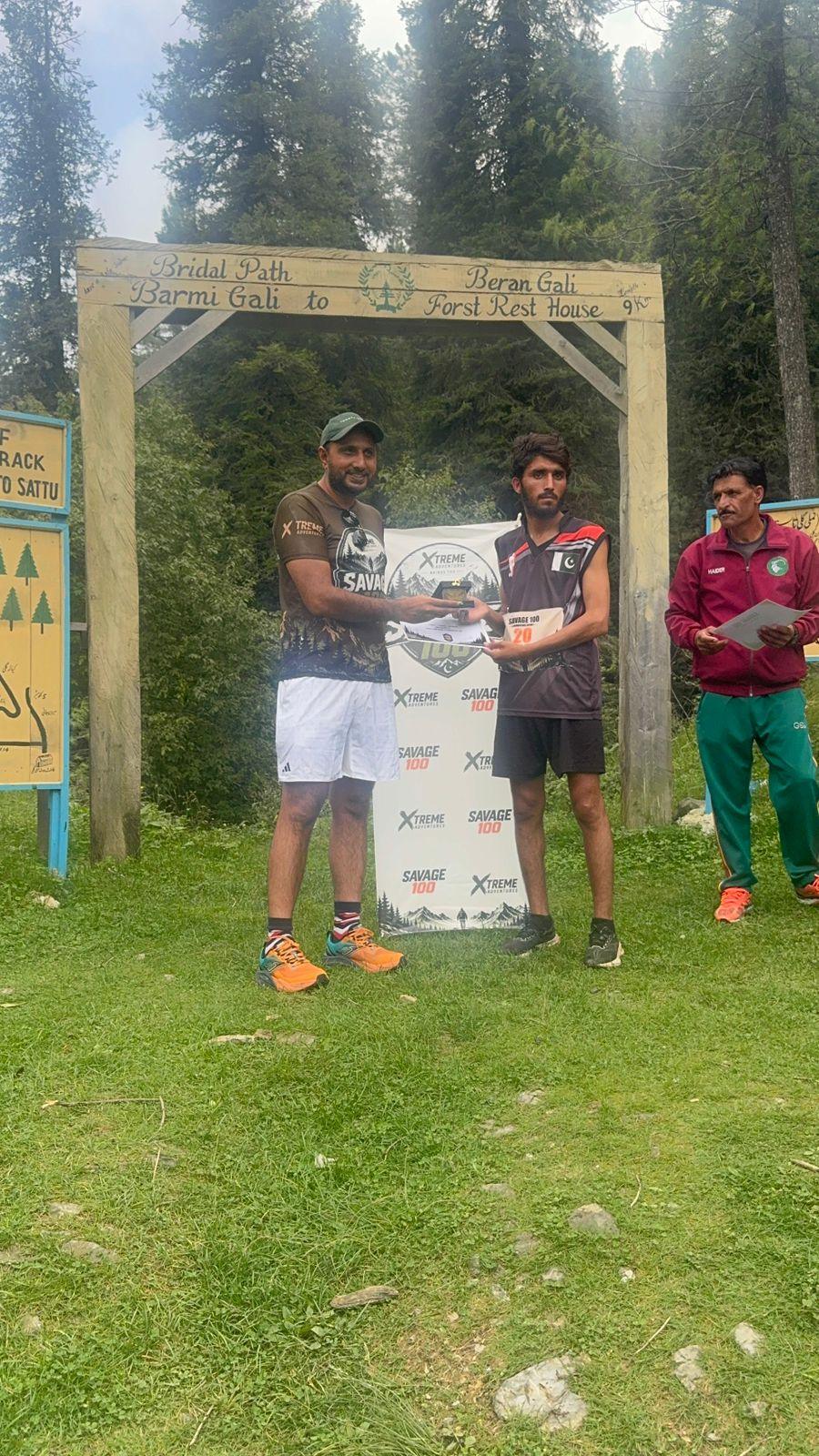 A slim runner in a maroon vest and dark shorts taking a certificate from the organiser under the wooden gate