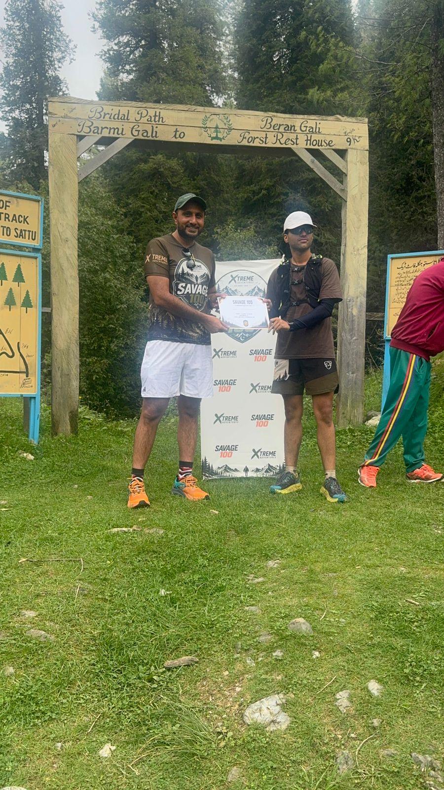 A finisher in a white cap, grey shirt and black shorts receiving a certificate under the gate beside the painted forest-track signboard