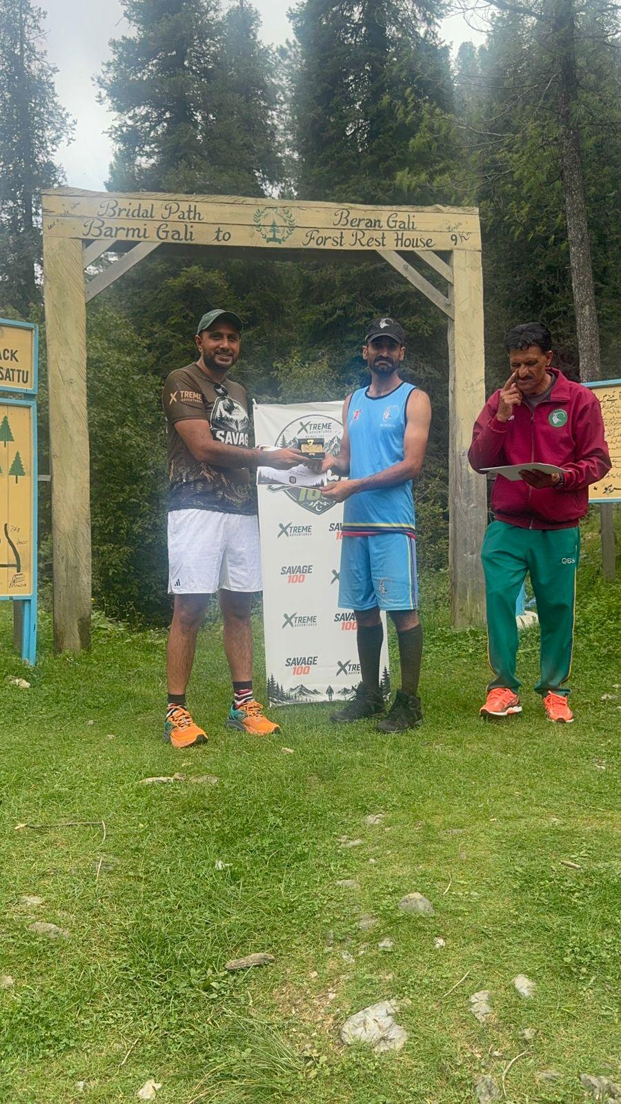 A finisher in a blue vest, blue shorts and rubber boots taking a certificate from the organiser under the wooden gate