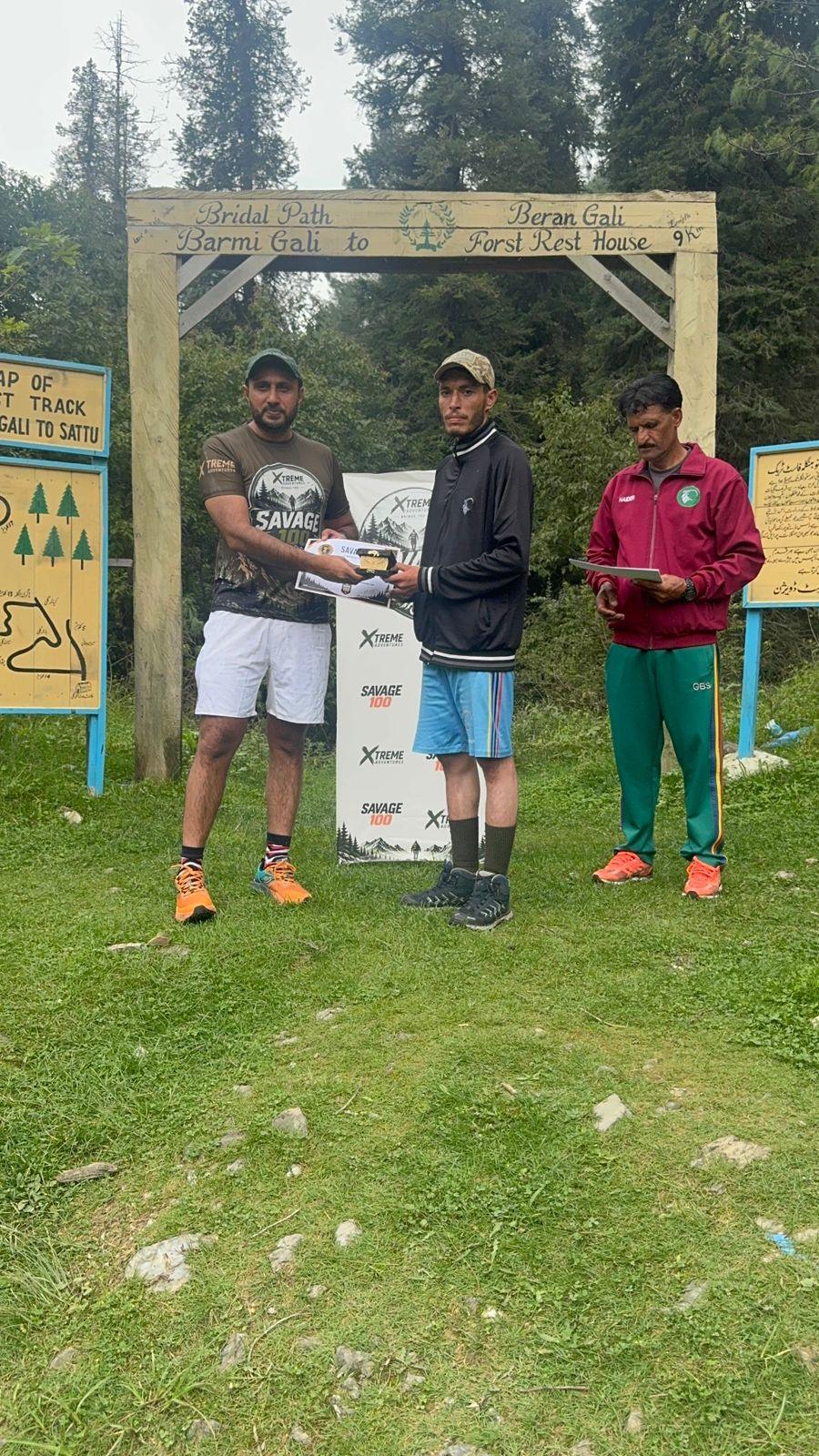 A finisher in a dark hooded top, cap and blue shorts receiving a certificate and a small trophy under the gate
