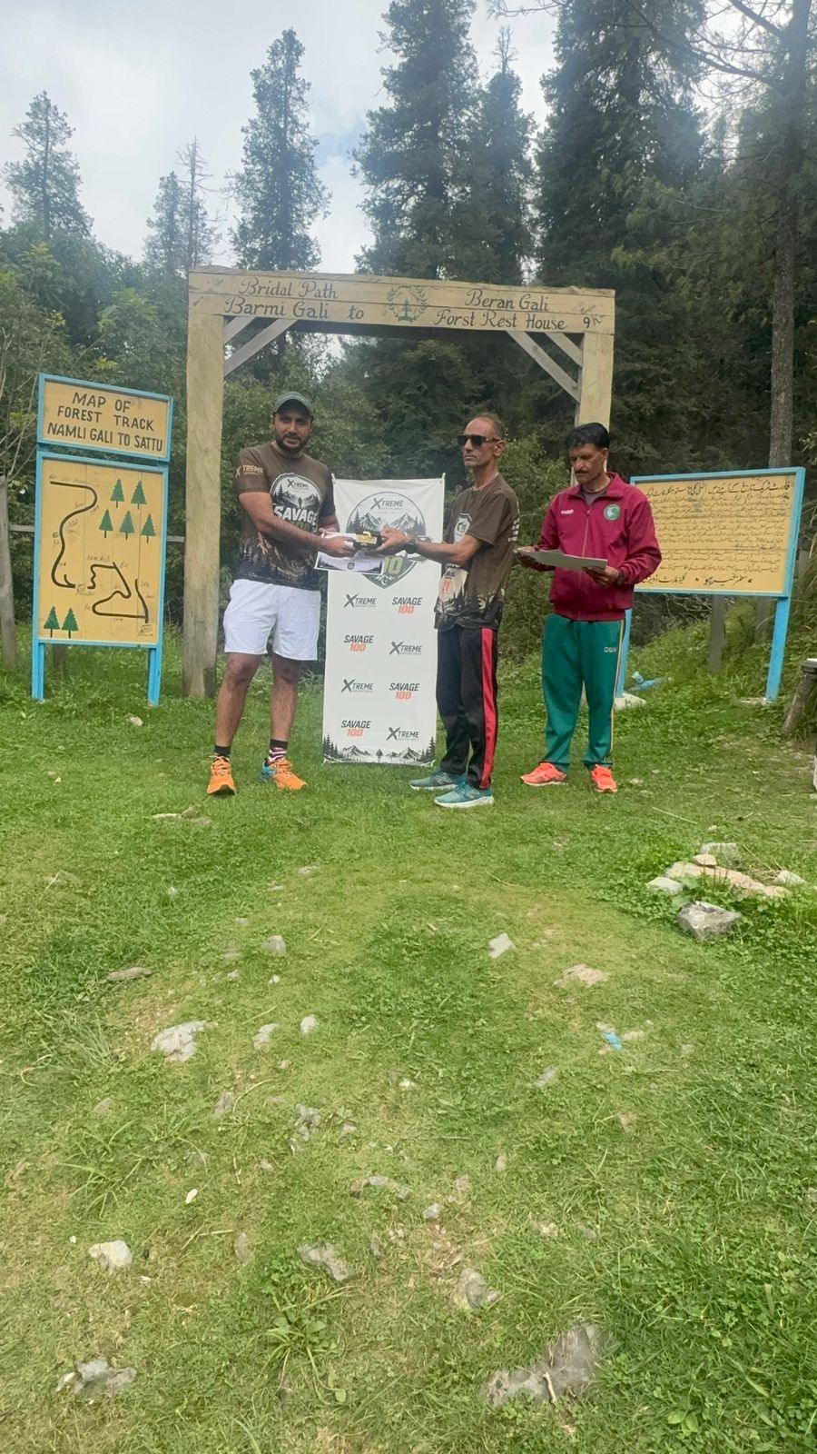A finisher in a grey shirt and dark trousers taking a certificate from the organiser, the marshal reading from a clipboard beside them