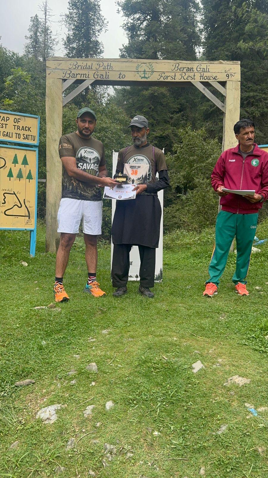 A finisher in a long black robe and cap receiving a certificate under the wooden gate in heavy cloud
