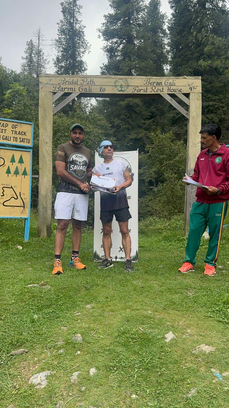 A finisher in a white shirt and grey shorts taking a certificate from the organiser under the gate, painted trail maps on the boards behind