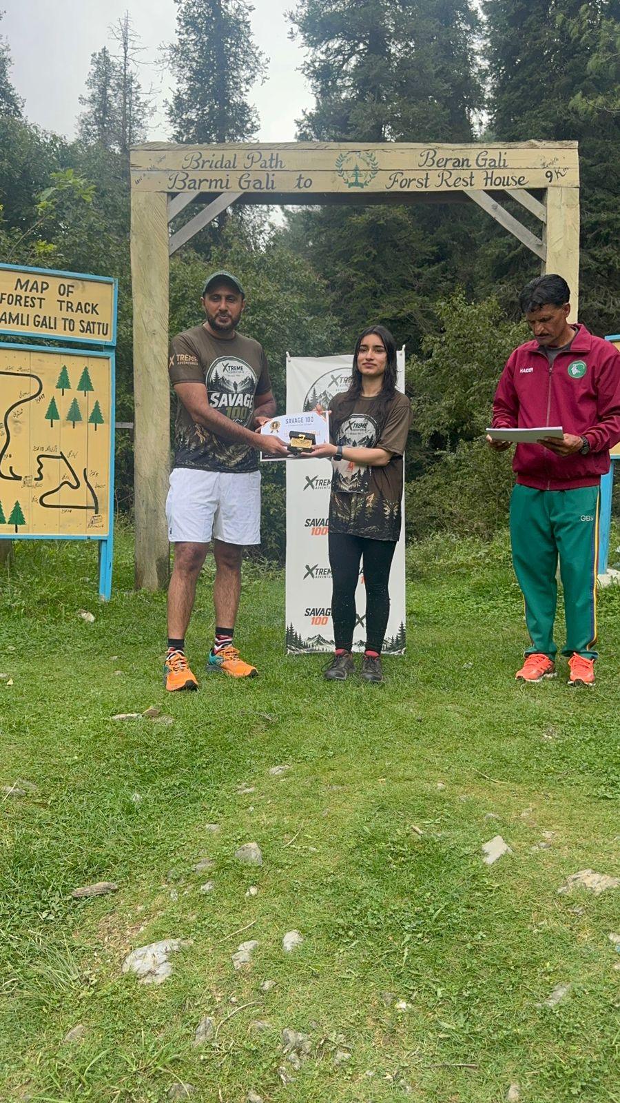 A woman in a dark patterned top receiving a certificate from the organiser under the wooden gate, the marshal with the clipboard to one side