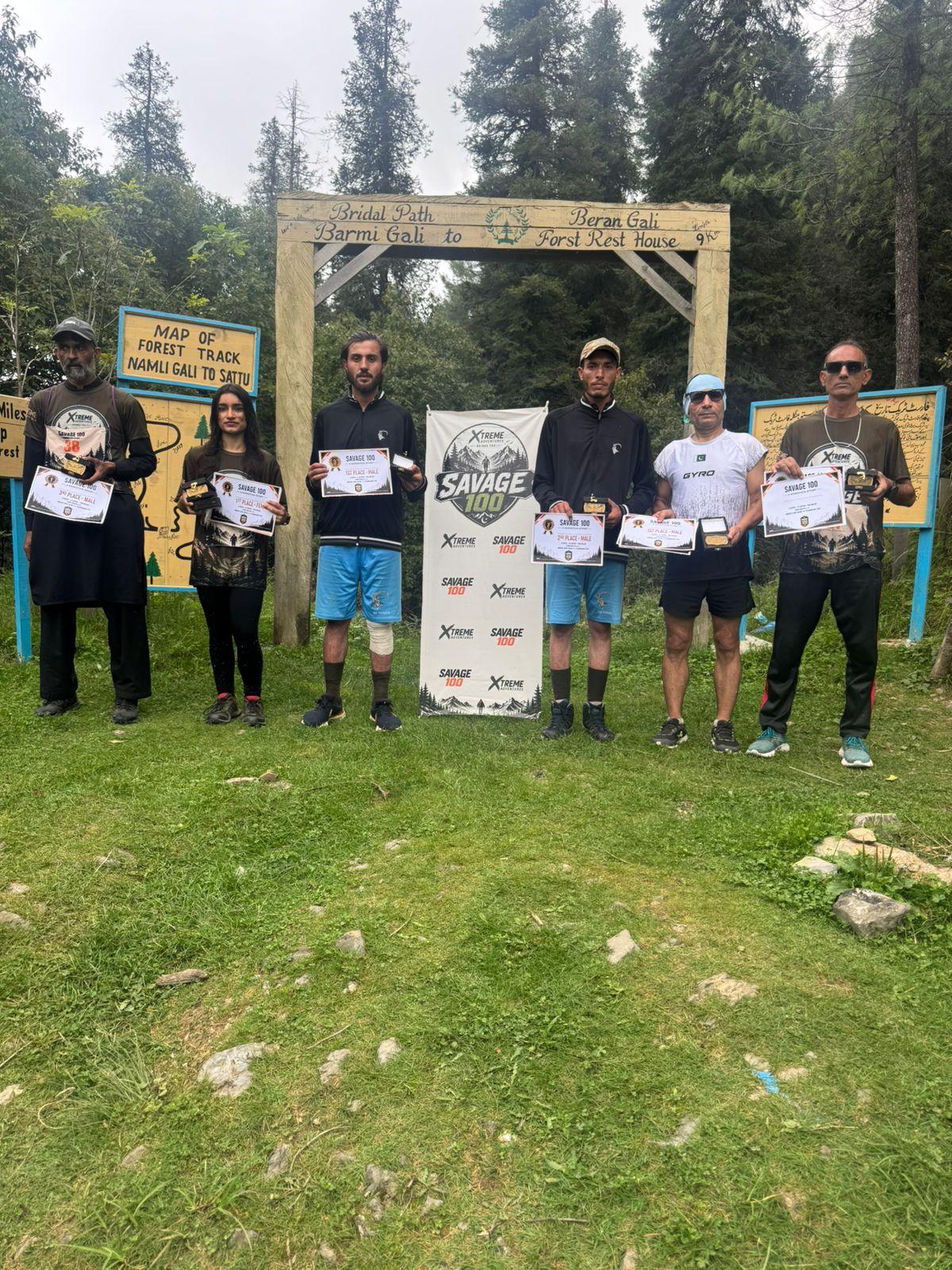 Six award winners standing in a line under the Beran Gali gate, each holding a certificate and a small trophy, the Savage 100 banner between them and painted forest-track signboards on either side